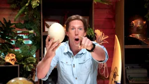 Een foto van Freek Vonk die een groot struisvogel ei in zijn hand heeft en geschrokken naar de camera wijst. 