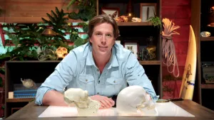 Een foto van Freek Vonk die een mensenschedel en een chimpansee schedel voor hem op tafel heeft liggen. 