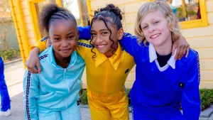Foto van drie kinderen in gekleurde outfits met armen om elkaars schouder. 