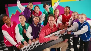 Foto van zangcoach met kinderen van koor in dezelfde sportieve jasjes met een keyboard.