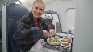 Een foto van presentator Janouk in een vliegtuig. Voor haar staan drie schaaltjes met eten. Daarnaast staat een flesje met water. Janouk kijkt in de camera.