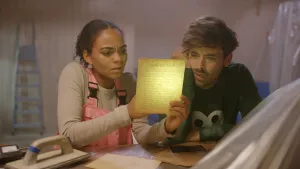 Een foto van de presentatoren Dzifa en Willem in een kamer. Ze zitten aan een tafel. Dzifa houdt een geel vel papier vast en schijnt er met een lampje op. Beiden kijken aandachtig naar de tekst op dat blaadje.