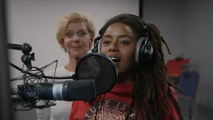 Een foto van presentator Eva. Ze staat in een opnamestudio achter een microfoon en heeft een koptelefoon op. Achter haar staat een vrouw die meekijkt.