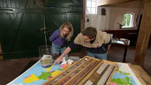 Een foto van presentatrice Janouk en een man bij een tafel. Op tafel ligt een landkaart van Europa en er liggen verschillende stenen. Er staat ook een glazen vaas met verschillende lagen aarde. De man wijst naar de verschillende lagen aarde.