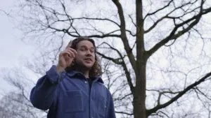 Een foto van een presentator in de buitenlucht. Hij staat voor een boom en wijst met zijn vinger in de lucht.