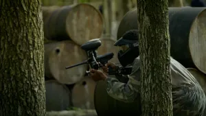 Foto van een persoon met helm en paintballgeweer die hurkt tussen de bomen.