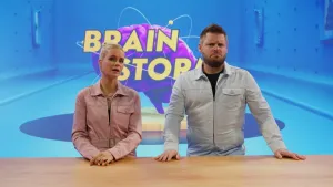 Foto van presentatoren Janouk en Job die naast elkaar staan en de camera inkijken. Janouk legt uit en Job kijkt moeilijk en vertwijfeld.