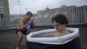 Een foto van een man die in een ijsbad zit, op de achtergrond zie je een man in zwembroek die squats doet.