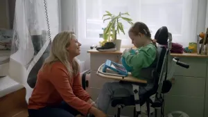 Een foto van een moeder, ze trekt schoenen aan van meisje met syndroom van Down die in een rolstoel zit.