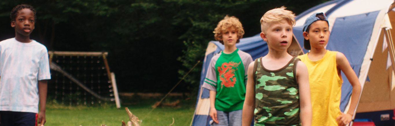 Een foto van kleuters op een cmaping, op de achtergrond zie je een tent en tussen hen in ligt een stapel takken.