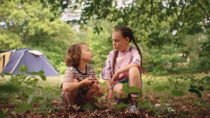 Een foto van twee kinderen op de camping, ze zitten op hun hurken onder een boom, op de achtergrond zie je een blauw tentje.