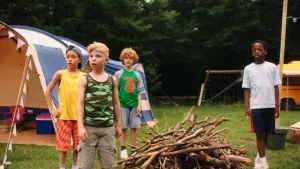Vier jongens op een camping, tussen hen in ligt een stapel takken en op de achterrond staat een tent.