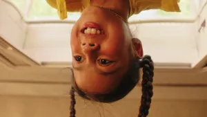 Een foto van een meisje dat ondersteboven hangt, je ziet alleen haar gezicht, haar vlechtjes hangen naar beneden.