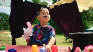 Een foto van een jongetje met een plaksnor op, een konijnenknuffel in zijn hand en wa bakjes op een tafel. Hij lijkt een goochelshow te geven.