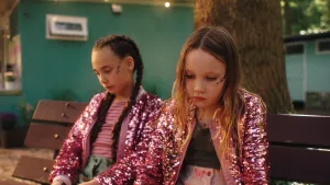 Twee meisjes in roze glitterjasjes zitten op een bankje en kijken sip.