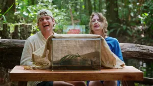 Een foto van de beide presentatoren, voor het staat een grote glazen bak waarover een doek zit. Jurre moet lachen, Emma heeft een soort verband/touw om haar neus en kin en hoofd.