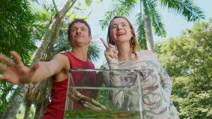 Een foto van de beide presentatoren bij een grote glazen bak die voor hen staat, ze kijken niet in de bak, maar schuin omhoog.