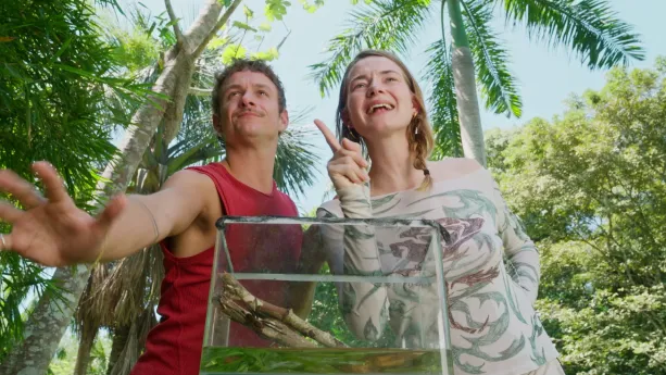 Een foto van de beide presentatoren bij een grote glazen bak die voor hen staat, ze kijken niet in de bak, maar schuin omhoog.