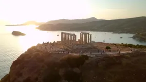 Een foto van een griekse tempel op een berg aan een baai, de zon  staat laag bij de ruine lopen allerlei mensen.