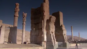 Een foto van de ruïnes van Persepolis in het oude Perzië, je ziet enkele pilaren, standbeelden en restanten van een muur tegen een strakblauwe lucht.