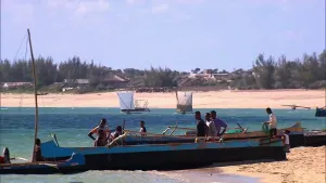 Een foto van mensen in houten boten bij de kust, po de achtergrond een zandstrand en huisjes en boten met een vierkant zeil.