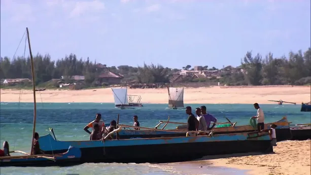Een foto van mensen in houten boten bij de kust, po de achtergrond een zandstrand en huisjes en boten met een vierkant zeil.