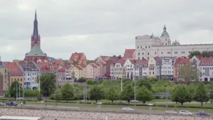 Een foto van een stadbeeld, je ziet een kade met daarachter een kerk, geel en wit gekleurde huizen met rode daken en een paar waar auto's voor langs rijden.