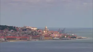 Een foto van een stadje aan de kust, de witte kerktoren licht op in de zon, het stadje steekt een stukje de zee in, je ziet huizen, kerken, kranen en linksachter een park of bos.