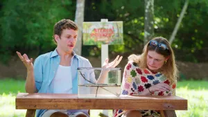 Een foto van de beide presentatoren, ze zitten biten aan een houten tafel waarom een bak staat. Jurre houdt zijn handen verbaasd omhoog en Emma kijkt griezelend. 