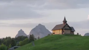Een foto van een Zwitsers landschap met een kerkje op een groene heuveltop, daarachter hoge grijze bergen.