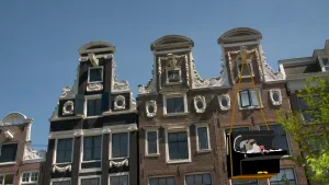 Een foto van grachtenhuizen in Amsterdam. Dit zijn hoge smalle huizen die mooi versierd zijn. Bij één van de huizen is een piano getekend die aan de hijsbalk hangt. Op de piano ligt een getekende duif te chillen.