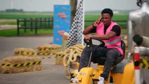 Foto van jongen die aan het bellen is terwijl hij een gele grasmaaier bestuurt op een parkours met hooibalen en andere obstakels. Hij kijkt nogal peinzend en heeft een hand aan het stuur.