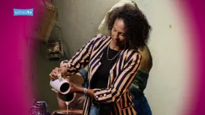 De presentatrice doet het melkmeisje na, achter haar is het schilderij te zien