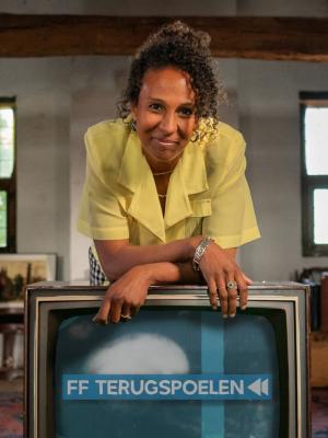 Presentatrice Astrid Sy staat op een oude, bruine zolder. Ze leunt over een televisie waar 'FF terugspoelen' in beeld staat.