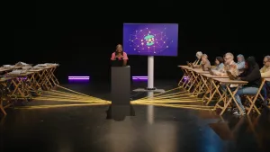 Een foto van presentatrice Tirsa die middenin een zaal achter een laptop staat. Links van haar tafels met boeken en kranten erop. Rechts van haar tafels met mensen achter laptops. Alle tafels zijn verbonden met waar zij staat door middel van gele touwen.