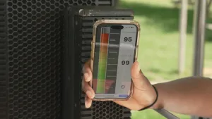 Een foto van een hand die een telefoon voor een geluidsbox houdt. Op het scherm van de telefoon is een decibelmeter te zien, die in het rood staat. Op het scherm staat 95 decibel.