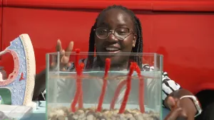 Een foto van presentatrice Tirsa die bij een glazen bak gevuld met water zit. In de bak staan rode draden recht overeind onder water. Links van haar is een stuk van een model van het oor te zien.