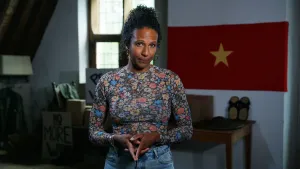 Presentatrice Astrid staat in het midden van het beeld. Ze staat op een zolder. Achter haar hangt een rode vlag met een ster erop.