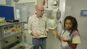 Een foto van presentatrice Eva die naast een man staat in een lab. Ze staan naast een machine  ten grootte van een flink koffiezetapparaat met een rijtje knoppen erop. De man houdt een bakje sla vast.
