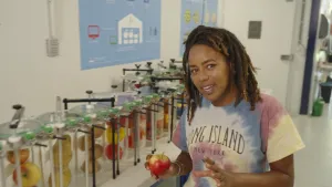 Een foto van presentatrice Eva met een appel in haar hand. Op de achtergrond staan hoge glazen bekers met appels, peren en paprika's erin.