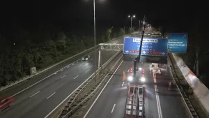 Een foto van een snelweg 's avonds in het donker. Op de snelweg staat een werkauto met een hijskraan. De kraan takelt een blauw bord met daarop plaatsnamen en de afstand omhoog. Op de weg lopen mensen in oranje veiligheidshesjes.