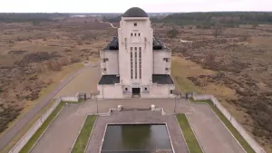 Een foto gemaakt met een drone van een hoog betonnen gebouw met kleine raampjes. het middelste gedeelte van het gebouw heeft een koepelvormig dak. Voor het gebouw ligt een vierkante vijver. Om het gebouw heen is alleen maar heide te zien.