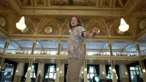 Een foto van presentator Pascal die in de hal van een hotel staat. Het dak is rijk versierd met schilderingen en er hangen grote luxe lampen: kroonluchters. Pascal heeft zijn camera uitgestrekt naar de camera alsof hij je uitnodigt mee te gaan.