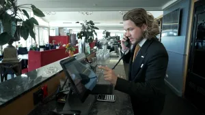 Een foto van presentator Pascal in een net zwart pak. Hij staat achter een bar in een hotel en heeft een telefoon aan zijn oor om een roomservice-bestelling op te nemen. Hij klikt iets aan op een beeldscherm.