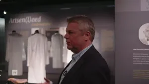 Een foto van een man, hij staat in een tentoonstelling, op de achtergrond zie je witte doktersjassen en de tekst Artsen(l)eed.