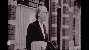 Een zwart-wit foto van gebedsgenezeres Greet Hofmans, ze staat buiten voor een gebouw met een jas over haar arm.