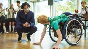 Jongen met groen shirt in roelstoel komt met zijn handen bij de grond. Een andere man zit gehurkt en spreekt met hem.