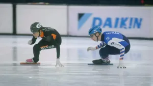 Een foto van twee schaatsers die snel door de bocht gaan op een schaatsbaan. Ze leunen beide in de bocht en houden hun hand op het ijs.