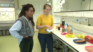 Presentatrice Eva en een vrouw staan in een laboratorium. De vrouw heeft een doos suikerklontjes in haar handen. Ze kijken naar een tafel met borden en glazen met verschillende soorten eten en drinken, zoals ontbijtkoek, druiven en chocolademelk.