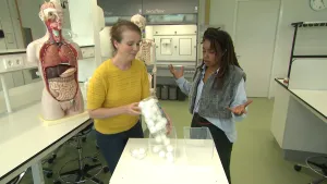 Een foto van presentatrice Eva en een vrouw in een laboratoriumruimte met witte tafels. De vrouw giet witte pingpongballetjes in één van de drie bakken op tafel. Op de achtergrond staat een model van het menselijk lichaam.